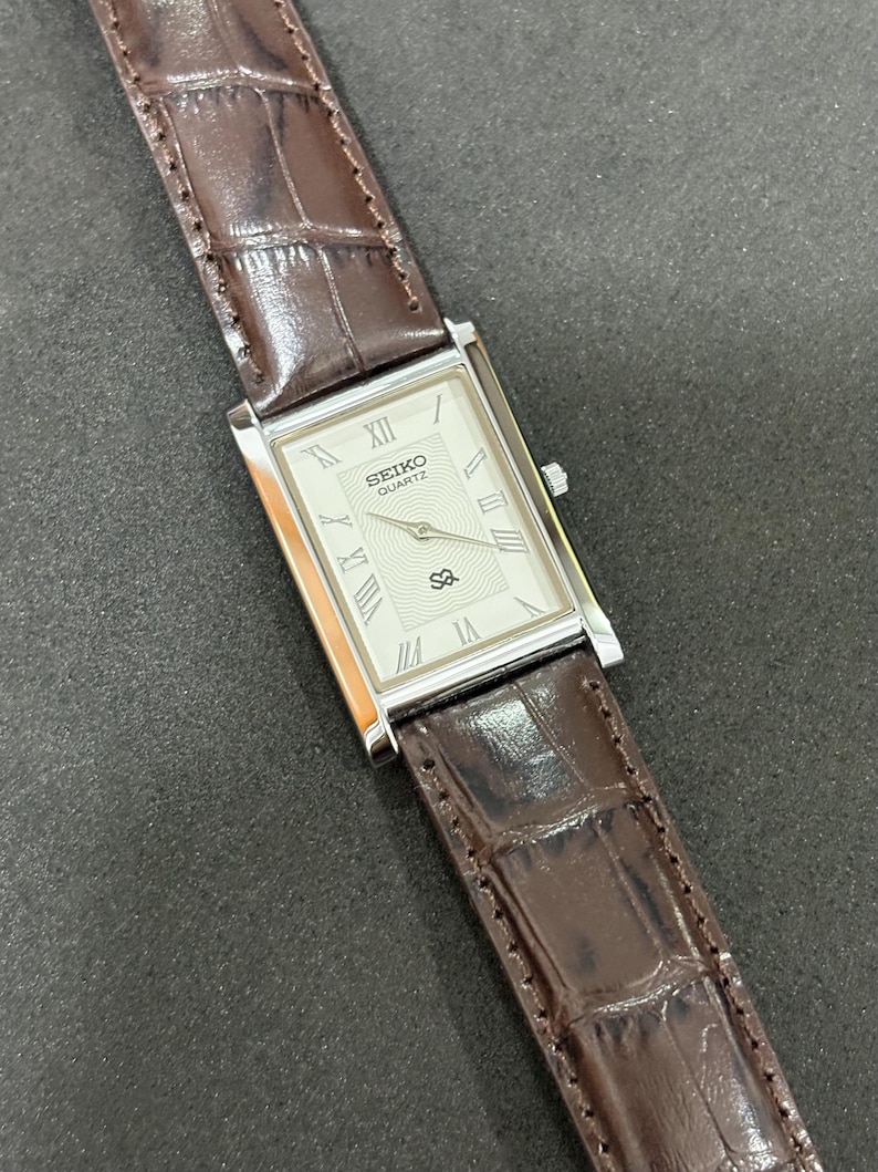 May include: A rectangular Seiko Quartz watch with a silver face and Roman numeral hour markers. It features a brown leather strap with a crocodile pattern. The watch face displays the Seiko Quartz logo.