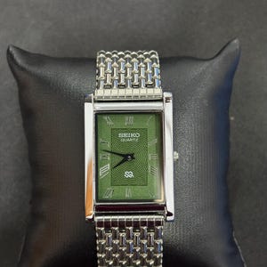 May include: A rectangular Seiko Quartz watch with a green face and silver-tone metal band. The watch face features Roman numerals and a textured design. The watch is displayed on a black pillow.