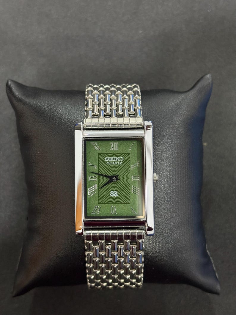 May include: A rectangular Seiko Quartz watch with a green face and silver-tone metal band. The watch face features Roman numerals and a textured design. The watch is displayed on a black pillow.