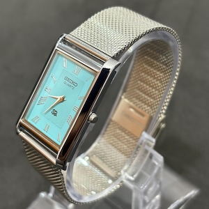 Può includere: Un orologio Seiko Quartz rettangolare con quadrante azzurro e cinturino in maglia d'argento. Il quadrante dell'orologio presenta numeri romani e il logo Seiko. L'orologio è esposto su un supporto trasparente.