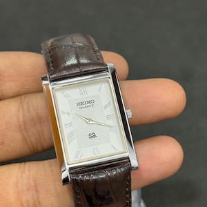 May include: A rectangular Seiko Quartz watch with a silver case and a white face with Roman numerals. The watch has a brown leather strap. The watch face displays the Seiko Quartz logo and a unique design element.