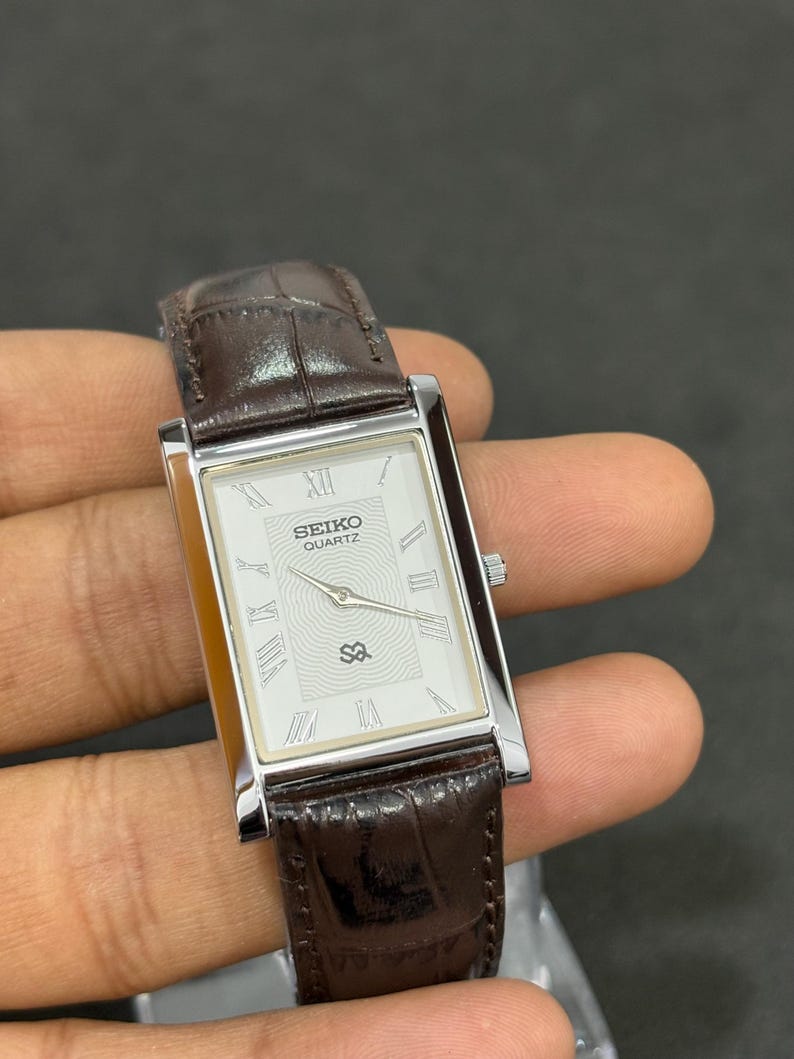May include: A rectangular Seiko Quartz watch with a silver case and a white face with Roman numerals. The watch has a brown leather strap. The watch face displays the Seiko Quartz logo and a unique design element.