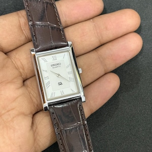 May include: A rectangular Seiko Quartz watch with a silver case and a white face featuring Roman numerals. The watch has a brown leather strap with a crocodile pattern. A classic wristwatch.