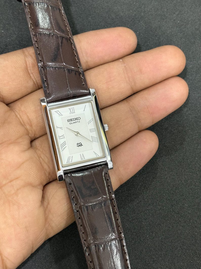 May include: A rectangular Seiko Quartz watch with a silver case and a white face featuring Roman numerals. The watch has a brown leather strap with a crocodile pattern. A classic wristwatch.