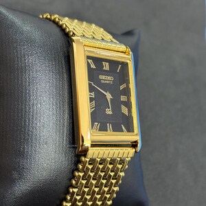 May include: A gold-toned Seiko Quartz watch with a rectangular black face and gold Roman numerals. The watch band is a woven gold mesh. The watch is displayed on a black surface.
