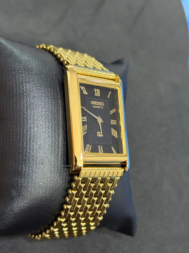 May include: A gold-toned Seiko Quartz watch with a rectangular black face and gold Roman numerals. The watch band is a woven gold mesh. The watch is displayed on a black surface.