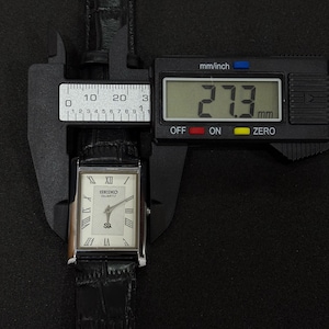 May include: A rectangular Seiko Quartz watch with a white face, Roman numeral hour markers, and a black leather band. The watch is measured at 27.3 mm using a digital caliper. The watch is displayed against a black background.