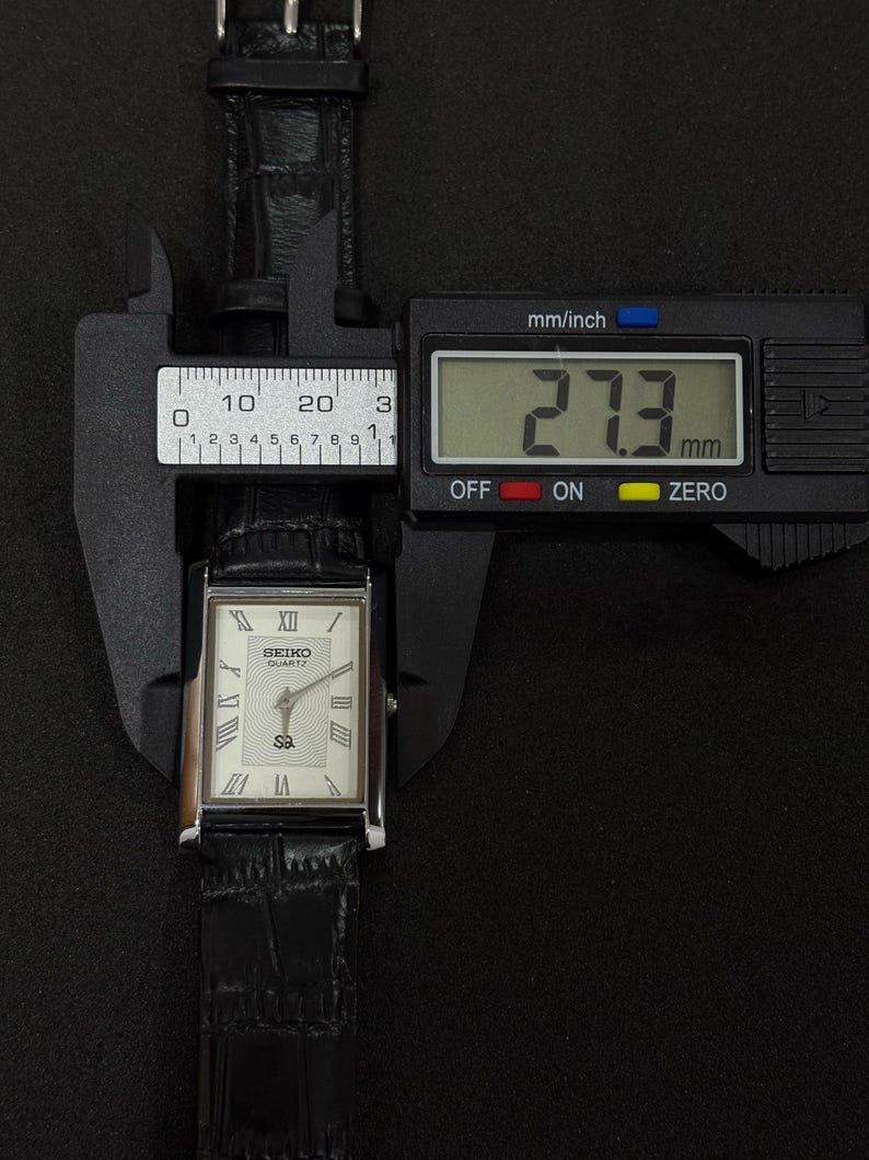 May include: A rectangular Seiko Quartz watch with a white face, Roman numeral hour markers, and a black leather band. The watch is measured at 27.3 mm using a digital caliper. The watch is displayed against a black background.