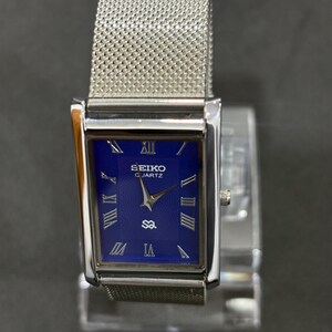 May include: A rectangular Seiko Quartz watch with a silver-tone case and mesh band. The watch face is a deep blue with silver Roman numerals and hands. The watch is displayed on a clear stand.