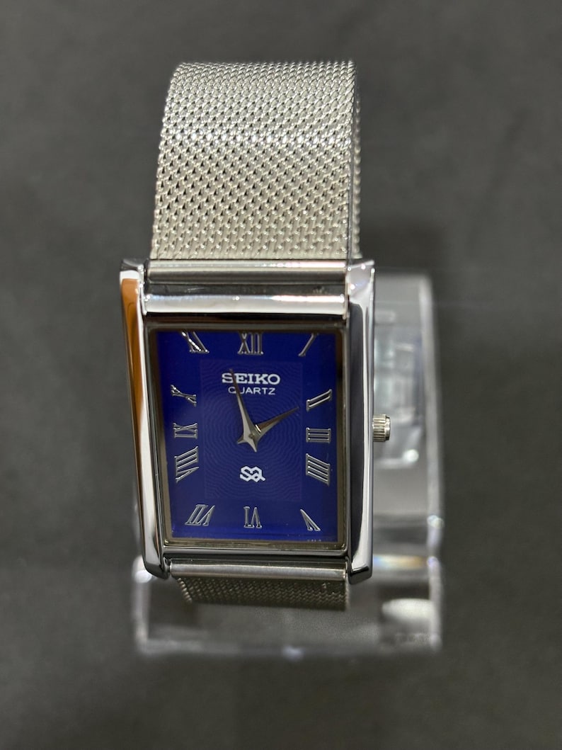 May include: A rectangular Seiko Quartz watch with a silver-tone case and mesh band. The watch face is a deep blue with silver Roman numerals and hands. The watch is displayed on a clear stand.