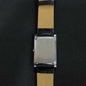 May include: A rectangular Seiko watch with a silver-colored back and a black leather band. The watch band is attached to the watch with a silver-colored metal piece. The watch band is black on the outside and tan on the inside.