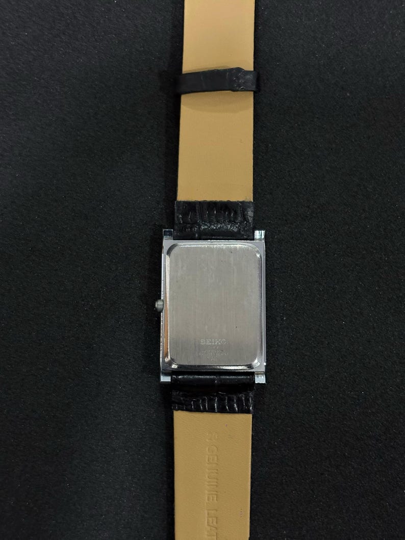 May include: A rectangular Seiko watch with a silver-colored back and a black leather band. The watch band is attached to the watch with a silver-colored metal piece. The watch band is black on the outside and tan on the inside.