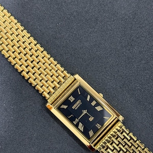 May include: A gold-toned Seiko Quartz watch with a rectangular black face and gold Roman numerals. The watch has a gold mesh band. The watch is angled on a dark gray surface.