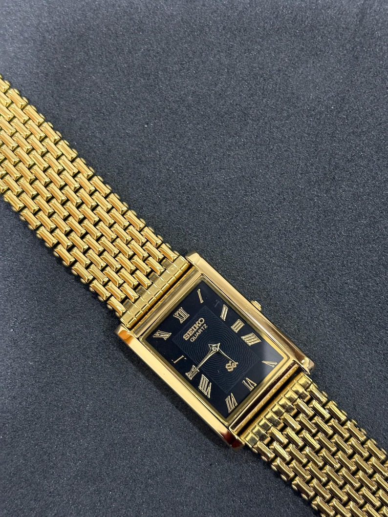 May include: A gold-toned Seiko Quartz watch with a rectangular black face and gold Roman numerals. The watch has a gold mesh band. The watch is angled on a dark gray surface.