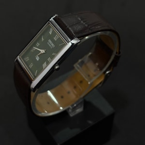 Może przedstawiać: Prostokątny zegarek Seiko Quartz z ciemnozieloną tarczą i srebrną kopertą. Zegarek posiada rzymskie cyfry i brązowy sk&oacute;rzany pasek. Zegarek jest prezentowany na czarnym stojaku.
