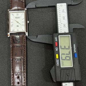 May include: A rectangular Seiko Quartz watch with a white face, Roman numeral hour markers, and a brown leather strap. The watch is being measured with a digital caliper, showing a width of 3.79 cm.