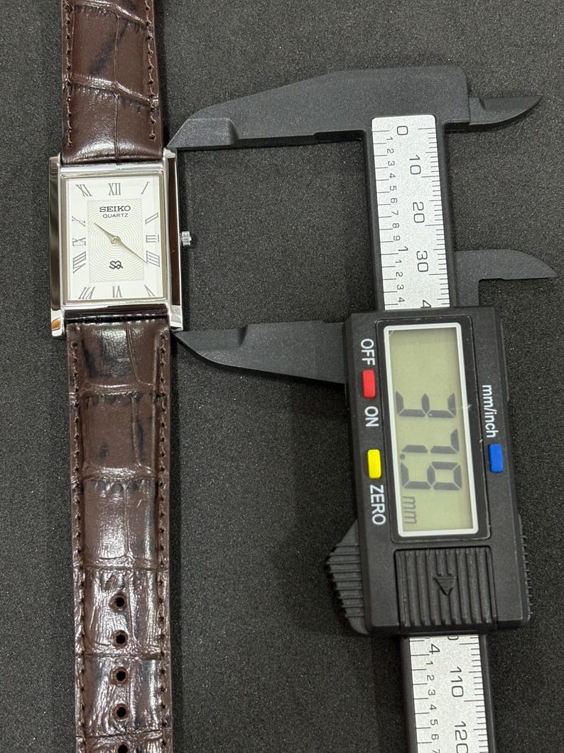 May include: A rectangular Seiko Quartz watch with a white face, Roman numeral hour markers, and a brown leather strap. The watch is being measured with a digital caliper, showing a width of 3.79 cm.