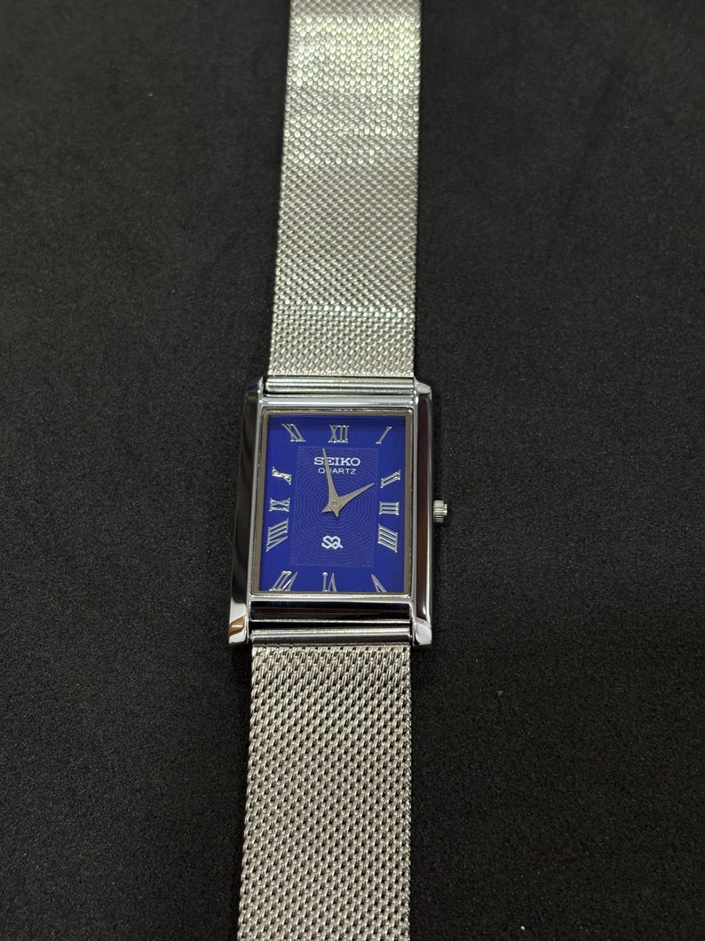 May include: A rectangular Seiko Quartz watch with a blue face and silver mesh band. The watch face features Roman numerals and the Seiko logo. The watch is displayed against a dark background.