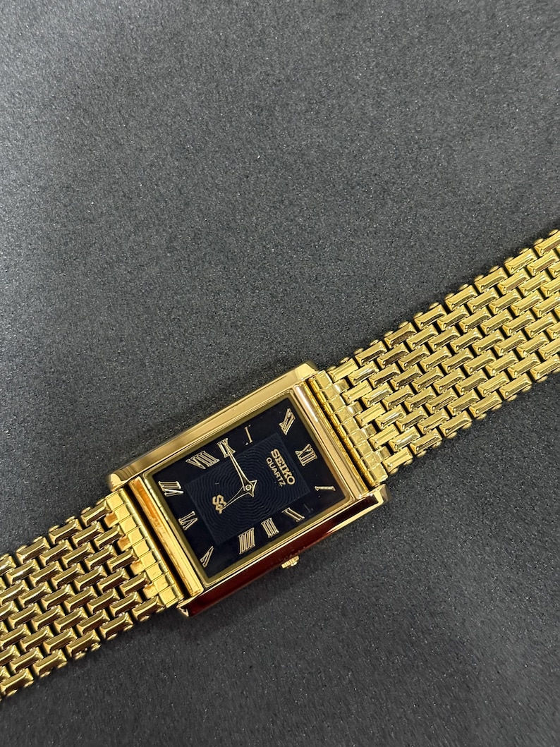 May include: A gold-toned Seiko Quartz watch with a rectangular black face and gold Roman numeral hour markers. The watch has a gold mesh band. The watch face displays the Seiko Quartz logo.