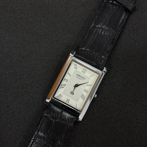 May include: A rectangular Seiko Quartz watch with a silver case and a white face featuring Roman numerals. The watch has a black leather band with a crocodile pattern. The watch face displays the Seiko Quartz logo.