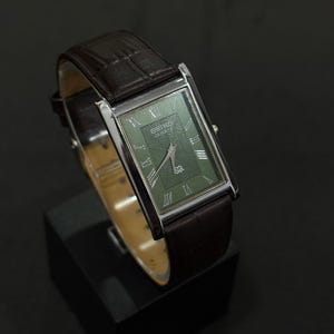 Może przedstawiać: Prostokątny zegarek Seiko Quartz ze srebrną kopertą i ciemnozieloną tarczą. Zegarek posiada rzymskie cyfry i brązowy sk&oacute;rzany pasek. Zegarek jest prezentowany na czarnym stojaku.
