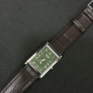 Może przedstawiać: Prostokątny zegarek Seiko Quartz z zieloną tarczą i srebrną kopertą. Zegarek posiada rzymskie cyfry jako znaczniki godzin i brązowy sk&oacute;rzany pasek. Zegarek jest prezentowany na ciemnej powierzchni.