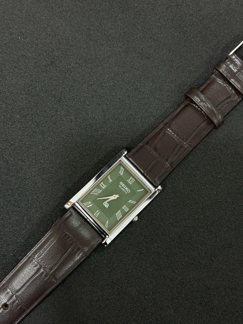 Może przedstawiać: Prostokątny zegarek Seiko Quartz z zieloną tarczą i srebrną kopertą. Zegarek posiada rzymskie cyfry jako znaczniki godzin i brązowy sk&oacute;rzany pasek. Zegarek jest prezentowany na ciemnej powierzchni.