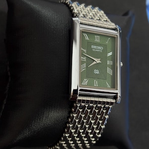 May include: A rectangular Seiko Quartz watch with a green face and silver-tone hands and Roman numeral hour markers. The watch has a silver-tone mesh band and is displayed on a black surface.