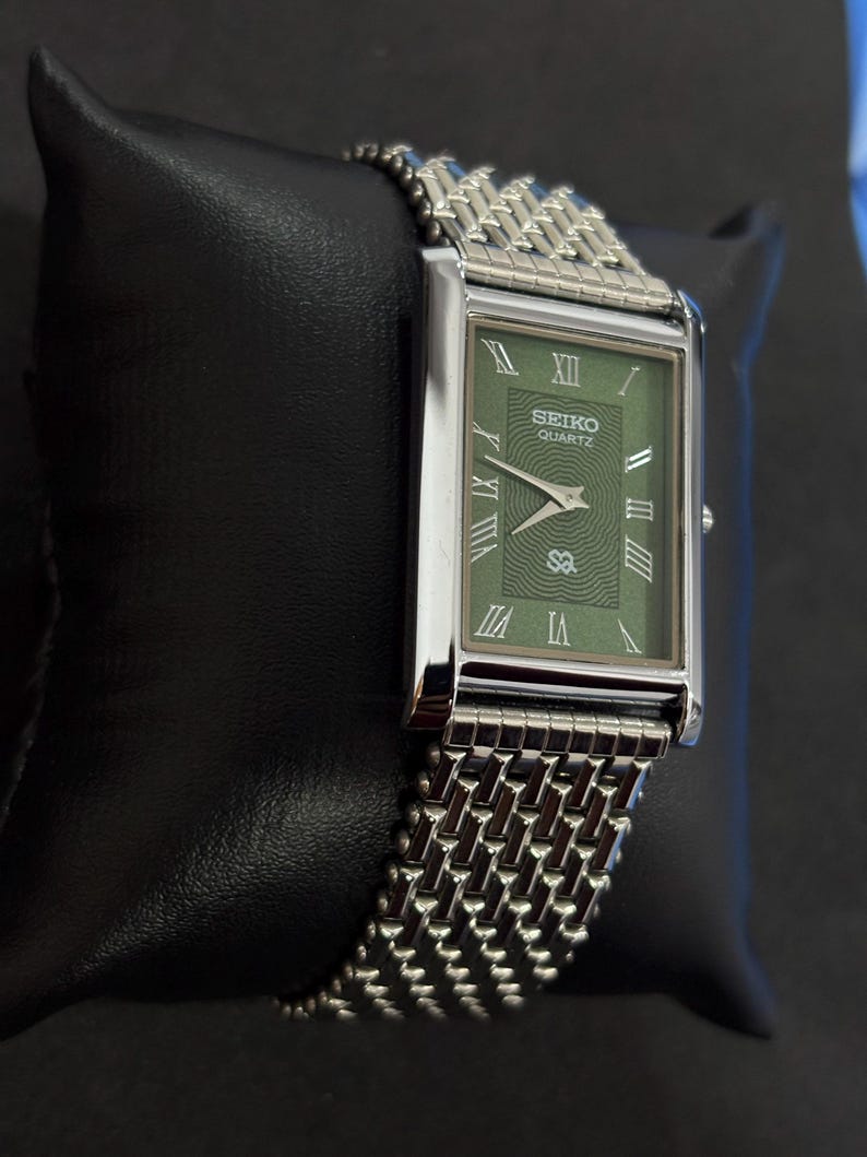 May include: A rectangular Seiko Quartz watch with a green face and silver-tone hands and Roman numeral hour markers. The watch has a silver-tone mesh band and is displayed on a black surface.