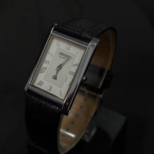 May include: A rectangular Seiko Quartz watch with a silver-tone case and a black leather band. The watch face is white with Roman numeral hour markers and a textured center. The watch is displayed on a black surface.