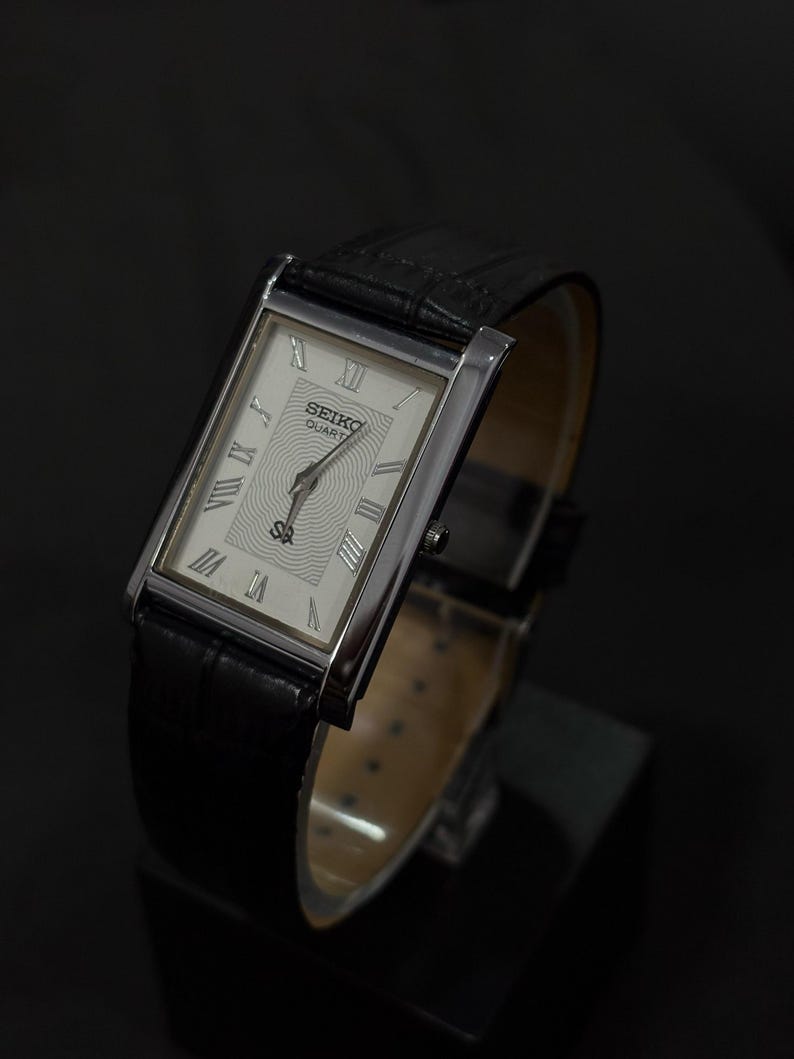 May include: A rectangular Seiko Quartz watch with a silver-tone case and a black leather band. The watch face is white with Roman numeral hour markers and a textured center. The watch is displayed on a black surface.