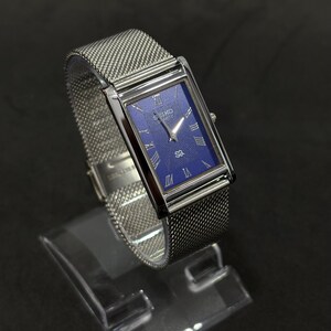 May include: A rectangular wristwatch with a silver mesh band and a blue face. The watch has silver Roman numeral hour markers and silver hands. The watch is displayed on a clear stand against a black background.
