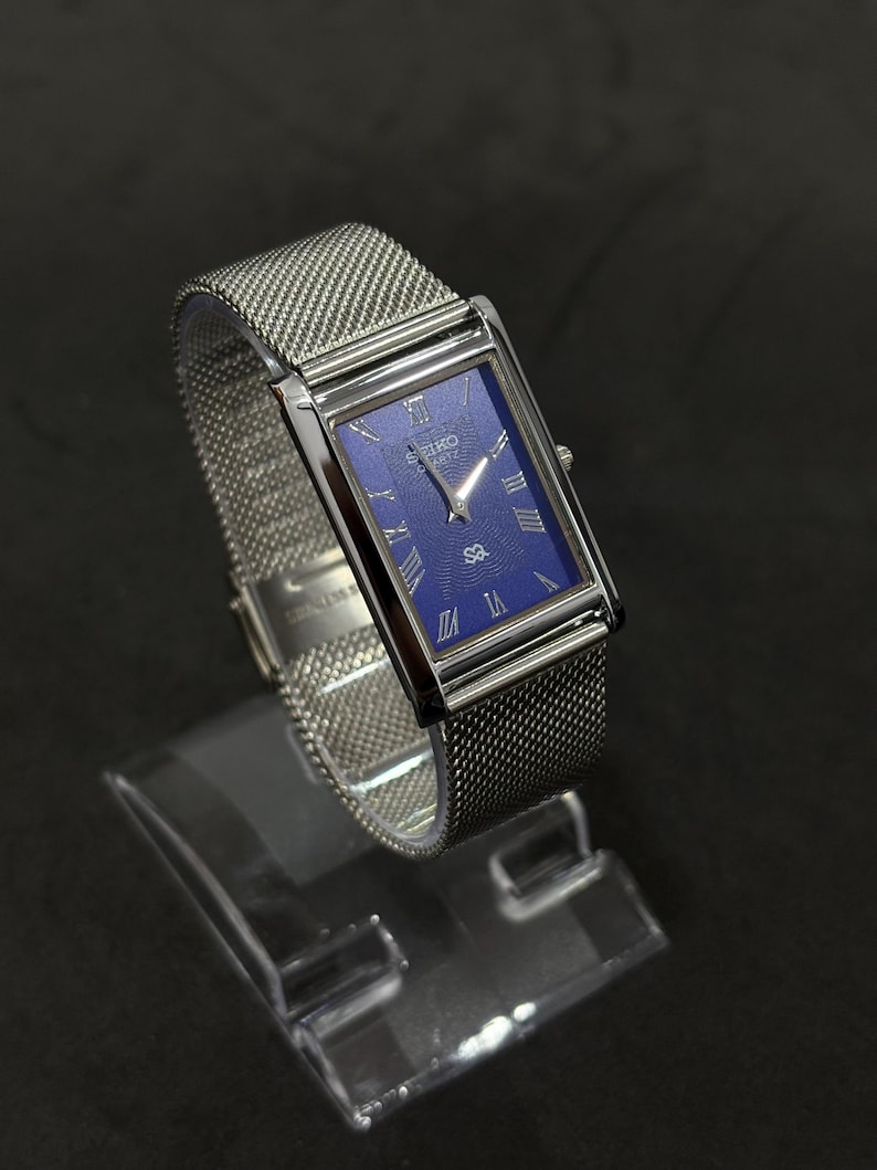 May include: A rectangular wristwatch with a silver mesh band and a blue face. The watch has silver Roman numeral hour markers and silver hands. The watch is displayed on a clear stand against a black background.