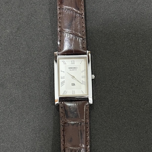 May include: A rectangular Seiko Quartz watch with a silver case and a white face with Roman numeral hour markers. It has a brown leather strap with a crocodile pattern. A classic watch design.