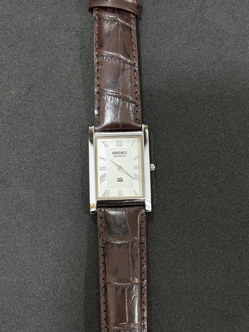 May include: A rectangular Seiko Quartz watch with a silver case and a white face with Roman numeral hour markers. It has a brown leather strap with a crocodile pattern. A classic watch design.