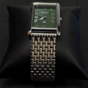 May include: A rectangular Seiko Quartz watch with a green face and silver-tone metal band. The watch face features Roman numerals and the Seiko logo. The watch is displayed on a black surface.