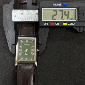 Może przedstawiać: Prostokątny zegarek Seiko Quartz z zieloną tarczą i rzymskimi cyframi. Brązowy sk&oacute;rzany pasek ma szerokość 27,4 mm. Zegarek jest mierzony suwmiarką cyfrową.