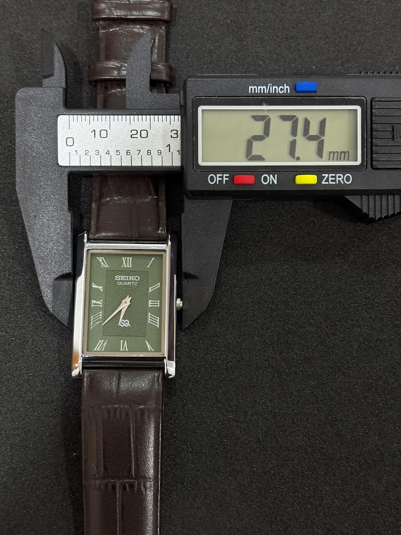 Może przedstawiać: Prostokątny zegarek Seiko Quartz z zieloną tarczą i rzymskimi cyframi. Brązowy sk&oacute;rzany pasek ma szerokość 27,4 mm. Zegarek jest mierzony suwmiarką cyfrową.