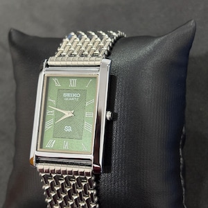 May include: A rectangular Seiko Quartz watch with a silver-tone case and a green dial featuring Roman numerals. The watch has a silver mesh band. The watch is displayed on a black pillow.