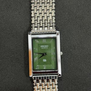 May include: A rectangular Seiko Quartz watch with a silver-tone metal band and case. The watch face is green with Roman numerals and the Seiko Quartz logo. The watch band has a woven design.