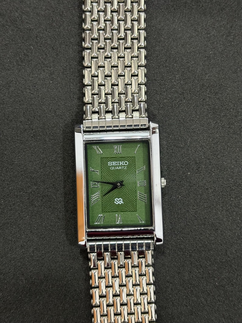 May include: A rectangular Seiko Quartz watch with a silver-tone metal band and case. The watch face is green with Roman numerals and the Seiko Quartz logo. The watch band has a woven design.