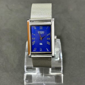 May include: A rectangular Seiko Quartz watch with a silver mesh band. The watch face is a vibrant blue with silver Roman numerals and hands. The watch is displayed on a clear stand.