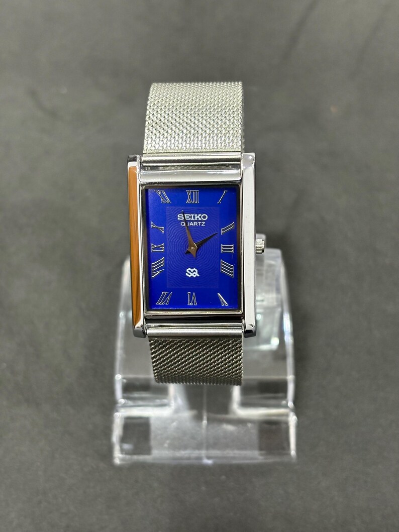 May include: A rectangular Seiko Quartz watch with a silver mesh band. The watch face is a vibrant blue with silver Roman numerals and hands. The watch is displayed on a clear stand.