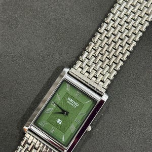 May include: A rectangular Seiko Quartz watch with a green face and silver-tone metal band. The watch face features Roman numerals and the Seiko logo. The band has a woven design.