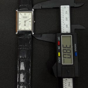 May include: A rectangular Seiko Quartz watch with a silver case and a black leather band. The watch face is white with Roman numeral hour markers. The watch measures 38.0 mm in width.