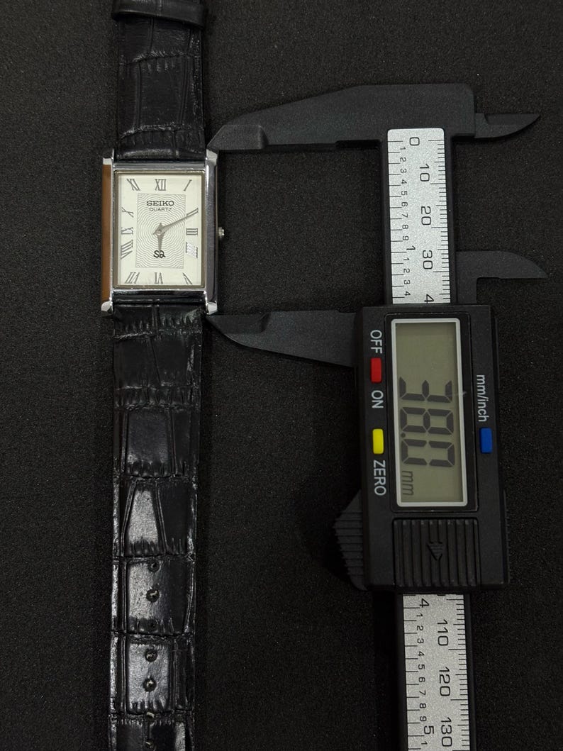 May include: A rectangular Seiko Quartz watch with a silver case and a black leather band. The watch face is white with Roman numeral hour markers. The watch measures 38.0 mm in width.