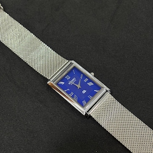 May include: A rectangular Seiko Quartz watch with a vibrant blue face and silver Roman numerals. The watch has a silver mesh band. The watch is displayed on a dark surface.