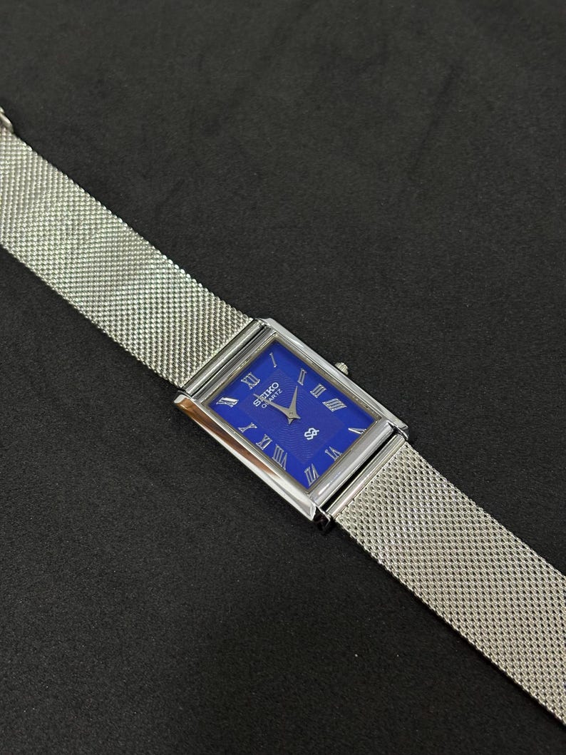 May include: A rectangular Seiko Quartz watch with a vibrant blue face and silver Roman numerals. The watch has a silver mesh band. The watch is displayed on a dark surface.