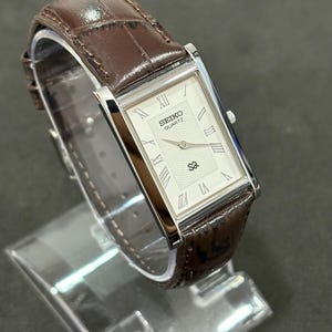 May include: A rectangular Seiko Quartz watch with a silver case and a cream-coloured face. The watch features Roman numeral hour markers and a brown leather strap with a crocodile pattern. The watch is displayed on a clear stand.