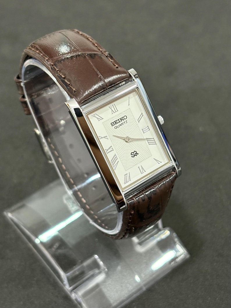 May include: A rectangular Seiko Quartz watch with a silver case and a cream-coloured face. The watch features Roman numeral hour markers and a brown leather strap with a crocodile pattern. The watch is displayed on a clear stand.
